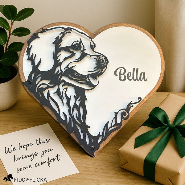 A heart-shaped wooden plaque with a custom black metal portrait of a Golden Retriever and the engraved name “Bella” sits on a light wooden surface. Beside it is a note reading, "We hope this brings you some comfort," and a gift wrapped in kraft paper with a green satin ribbon. A potted plant and soft neutral decor add warmth to the scene. The Fido & Flicka logo is in the corner.