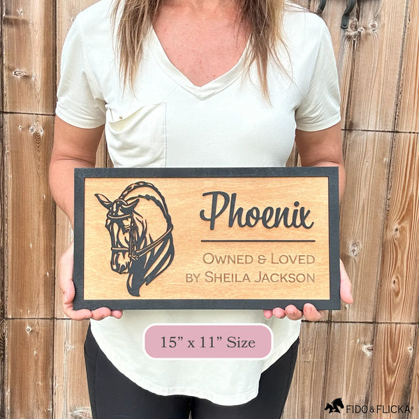 Woman holding custom wooden horse stall sign made from Baltic birch. Personalized plaque shows black dressage horse head design with engraved text horse name, Owned & Loved by owner. Sign measures 15” x 11”, rustic barn name plate.