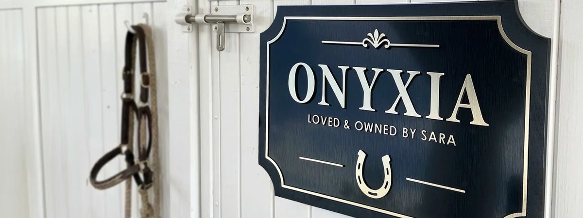  A close-up of a custom acrylic and wood horse stable door sign with the name 'ONYXIA' and the phrase 'LOVED & OWNED BY SARA' in white lettering on a dark navy plaque, mounted on a white painted wooden door next to a horse halter.