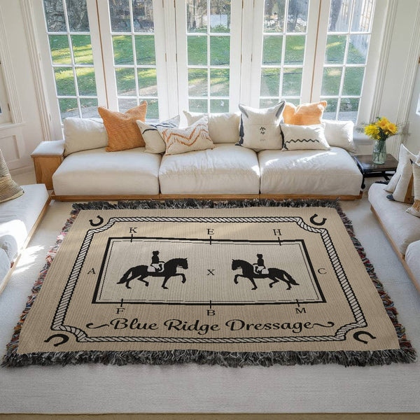 Large personalized woven cotton dressage arena blanket with rider silhouettes, rope border, and custom name displayed in a bright living room.