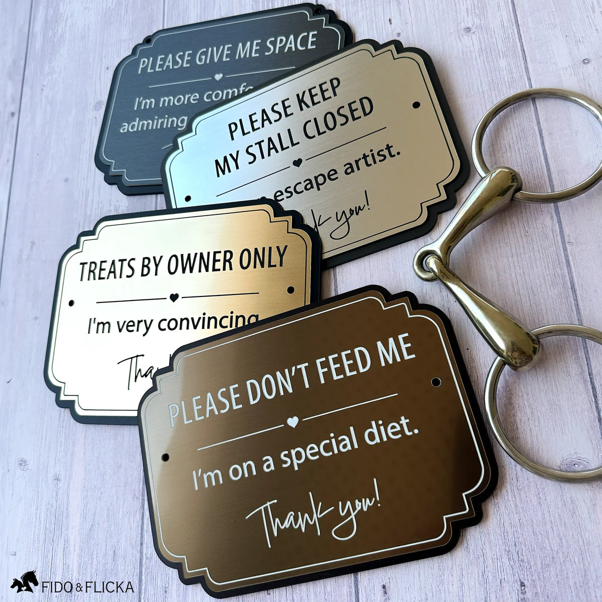 Four personalized horse stall warning signs in brushed metallic finishes displayed with an equestrian horse bit, showing different colors and messages.