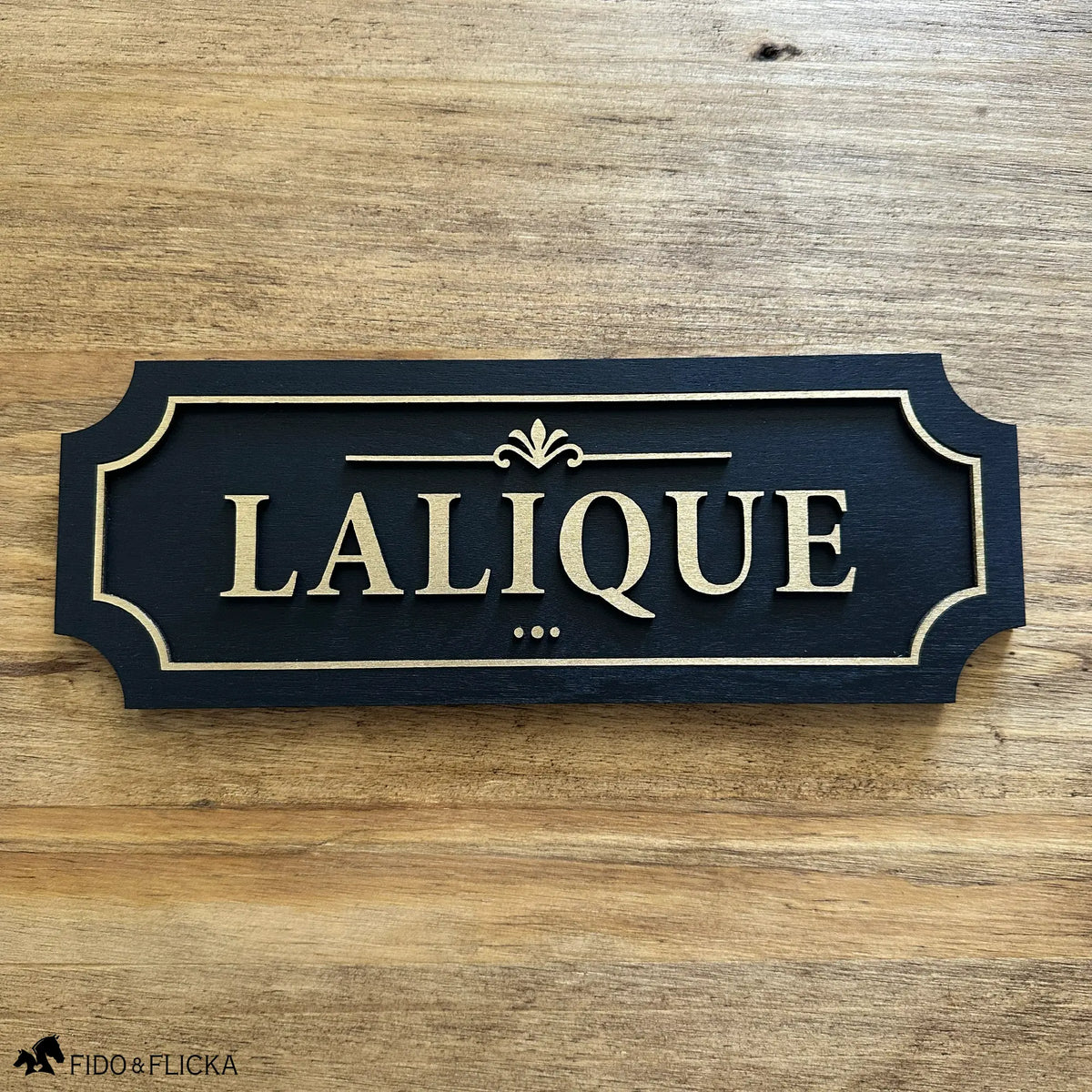 Black and gold layered wood horse stall name plate reading ‘Lalique,’ featuring raised gold lettering, an ornate gold accent, and a gold border on a black painted base, displayed on a natural wood surface.