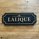 Black and gold layered wood horse stall name plate reading ‘Lalique,’ featuring raised gold lettering, an ornate gold accent, and a gold border on a black painted base, displayed on a natural wood surface.