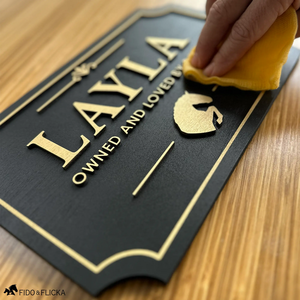 Hand wiping a black and gold layered wood horse stall name plate reading ‘Layla,’ featuring raised gold lettering and a gold horse silhouette on a wood surface.