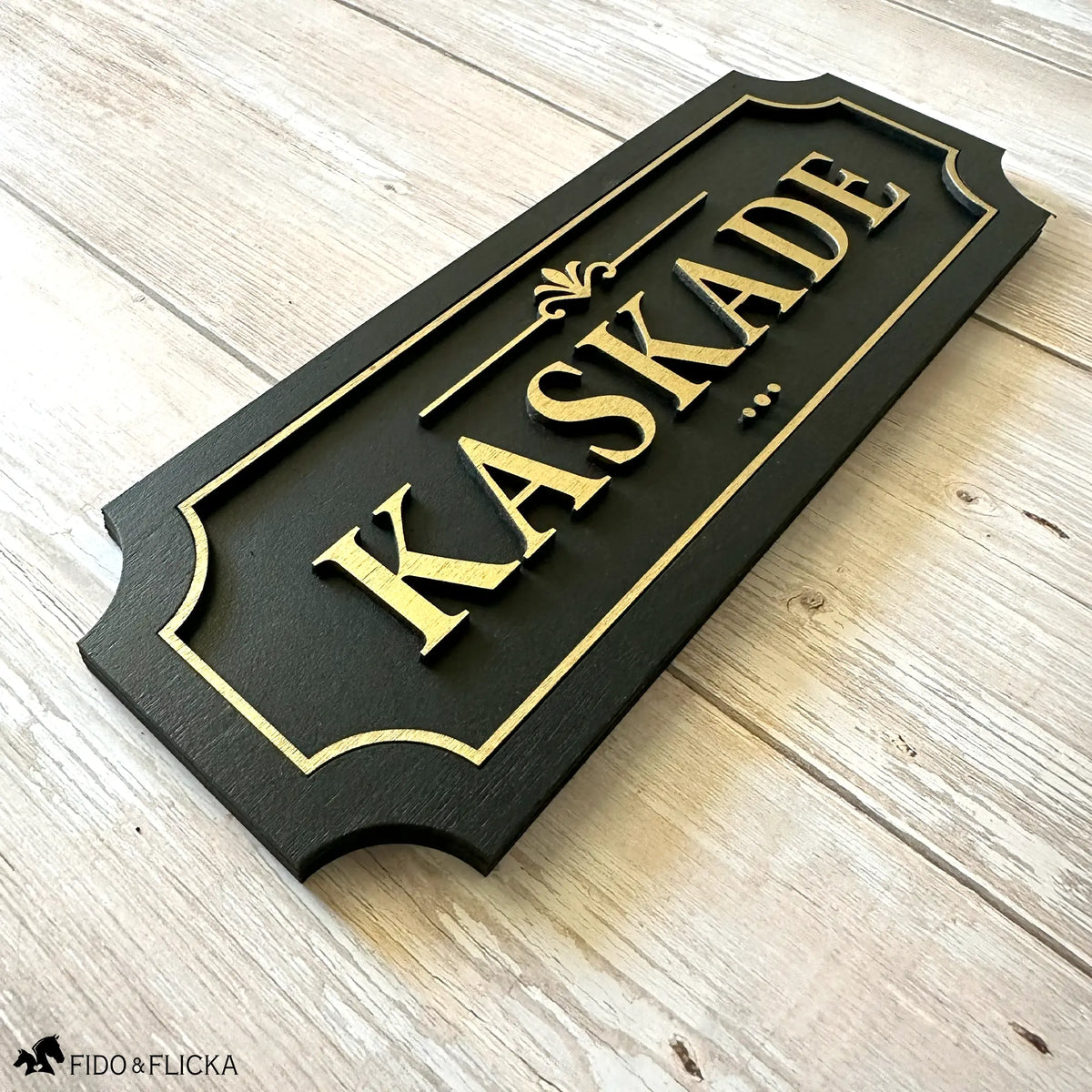 Black and gold layered wood horse stall name plate reading 'Kaskade,' featuring raised gold lettering and an elegant gold border on a black painted wood base, displayed on a light wood surface.