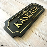 Black and gold layered wood horse stall name plate reading 'Kaskade,' featuring raised gold lettering and an elegant gold border on a black painted wood base, displayed on a light wood surface.