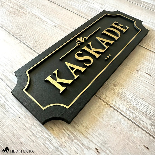 Black and gold layered wood horse stall name plate reading 'Kaskade,' featuring raised gold lettering and an elegant gold border on a black painted wood base, displayed on a light wood surface.
