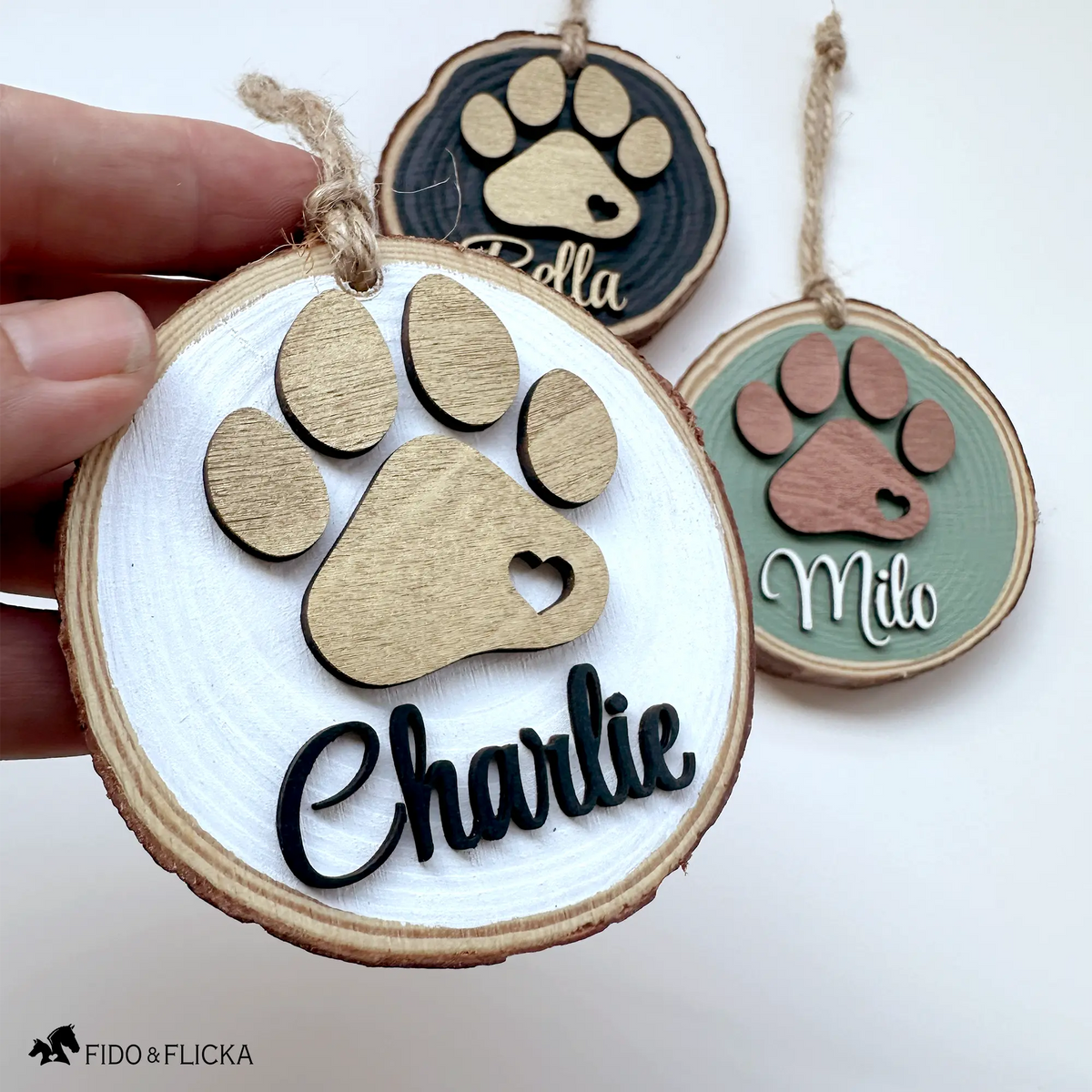 Hand holding a personalized wood slice dog ornament with a layered gold paw print and the name “Charlie” in black script, with additional sage green and black color options displayed in the background.