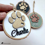 Hand holding a personalized wood slice dog ornament with a layered gold paw print and the name “Charlie” in black script, with additional sage green and black color options displayed in the background.