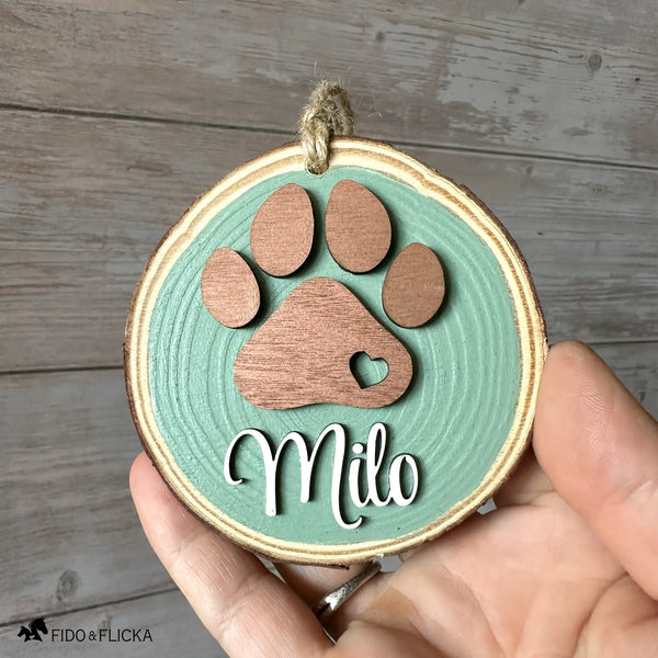Hand holding a personalized sage green wood slice dog ornament featuring a layered rose gold paw print with a heart cutout and the name “Milo” in white script, shown against a rustic wood background.