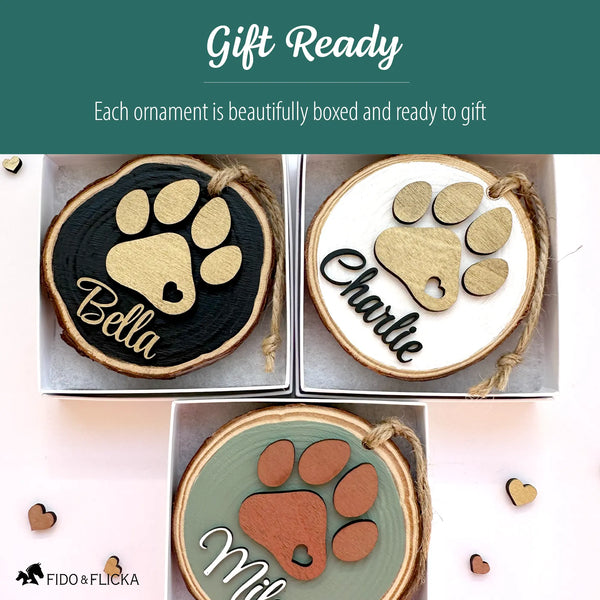 Three personalized dog paw wood slice ornaments—black, white, and sage—shown packaged in gift boxes, highlighting the gift-ready presentation and layered wooden paw prints with custom names.
