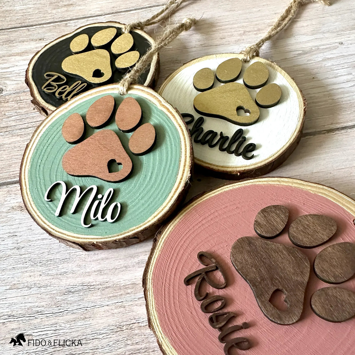 Four personalized wood slice dog ornaments displayed in a row, showing the color options: black with gold paw, white with gold paw, sage green with rose gold paw, and dusty pink with dark oak paw. Each ornament features a layered paw print and custom pet name.