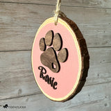 Side-angle view of a personalized dusty pink wood slice dog ornament with a layered dark oak paw print and the name “Rosie” in matching dark wood script, hanging by natural jute twine against a rustic wood wall.