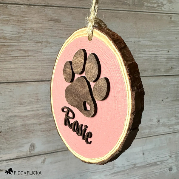 Side-angle view of a personalized dusty pink wood slice dog ornament with a layered dark oak paw print and the name “Rosie” in matching dark wood script, hanging by natural jute twine against a rustic wood wall.