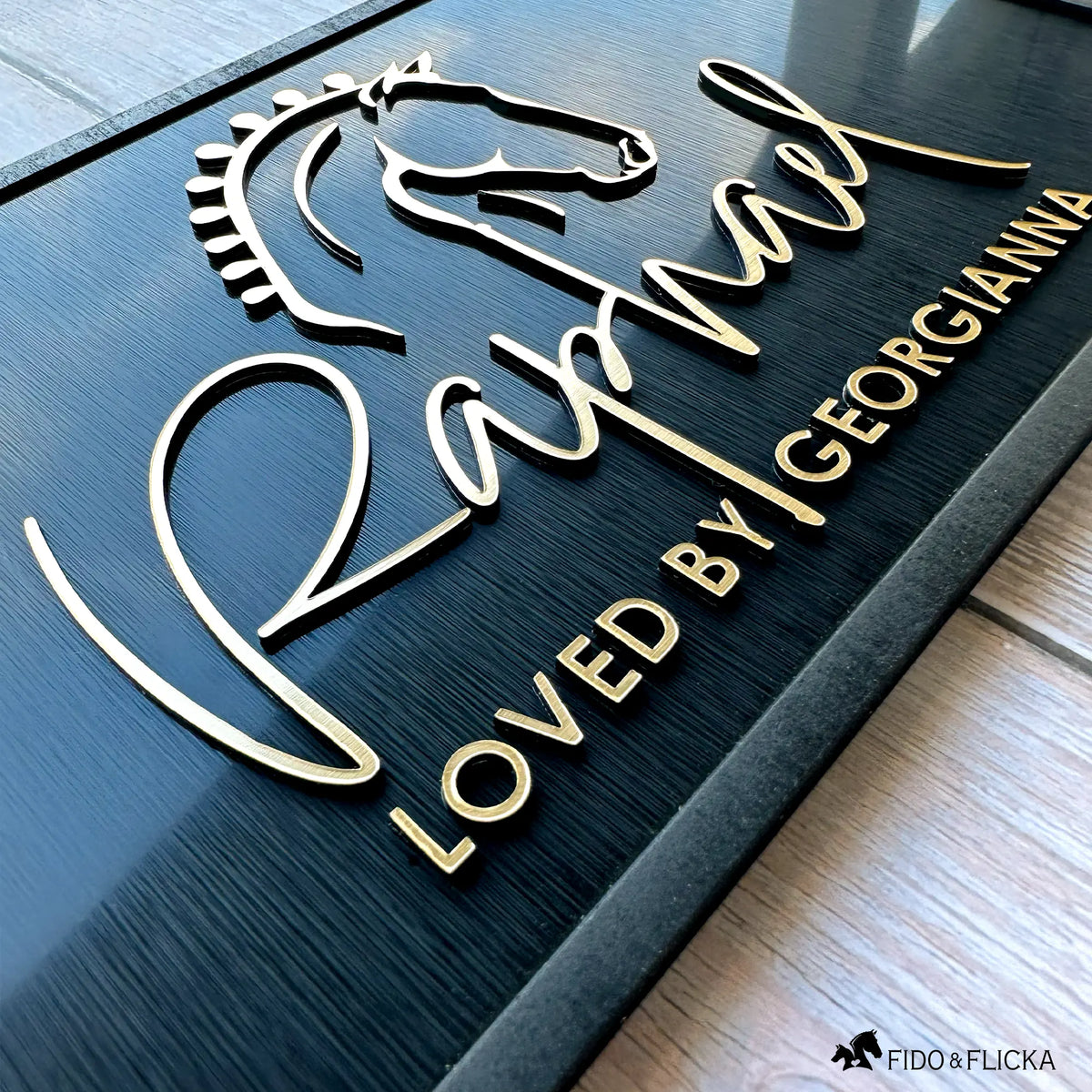 Close-up angled view of a custom horse stall nameplate by Fido & Flicka featuring brushed gold acrylic show horse silhouette and the name “Raphael” with the inscription “Loved by Georgianna.” The design is laser-cut with a matte black wood frame with elegant raised acrylic lettering, showcasing fine craftsmanship and modern equestrian style.