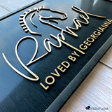 Close-up angled view of a custom horse stall nameplate by Fido & Flicka featuring brushed gold acrylic show horse silhouette and the name “Raphael” with the inscription “Loved by Georgianna.” The design is laser-cut with a matte black wood frame with elegant raised acrylic lettering, showcasing fine craftsmanship and modern equestrian style.