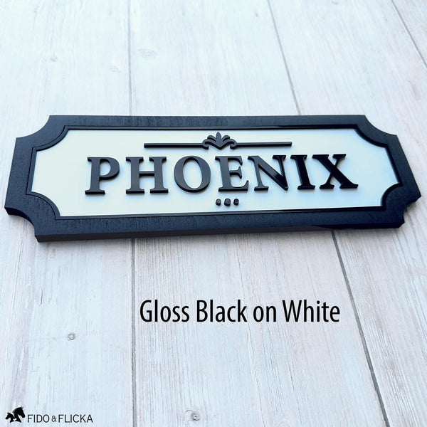 Custom horse stall name plate with raised gloss black acrylic lettering on a white background, framed in black wood and personalized with the name Phoenix.