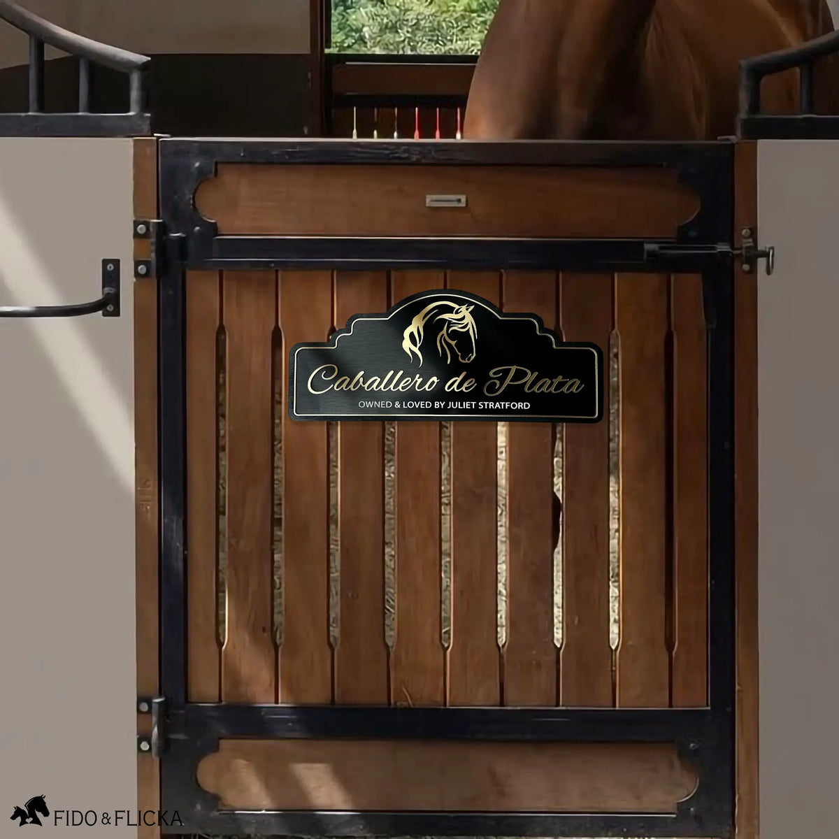 Personalized horse stall sign mounted on a wood and black iron stall door with a chestnut horse visible in the background. The arch-top sign reads "Caballero de Plata, Owned and Loved by Juliet Stratford" in raised brushed gold acrylic lettering on a black background with a gold border and horse portrait silhouette. Shown in context on a premium barn stall door.