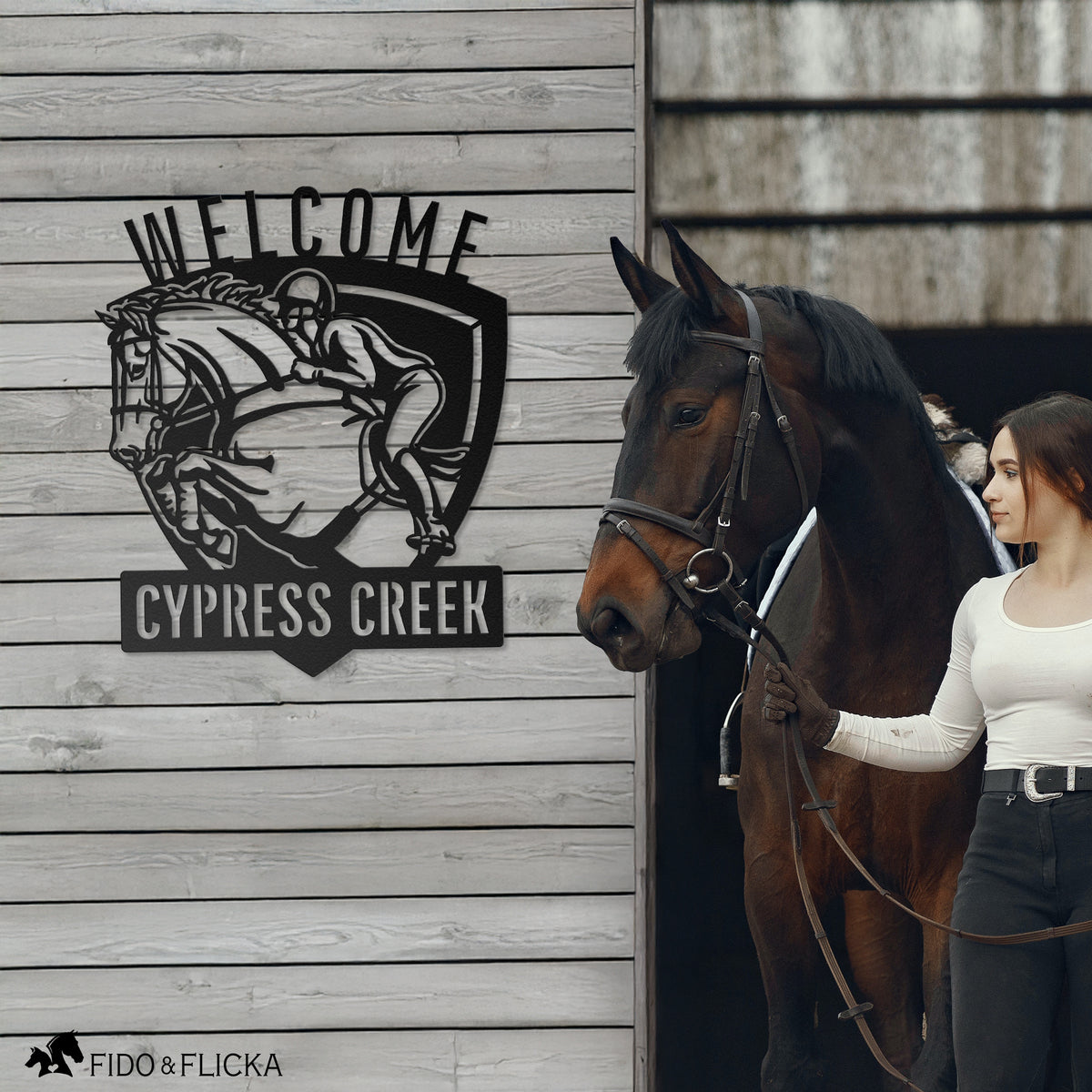 Personalized metal welcome sign for horse barn featuring show jumping horse and rider with custom text ‘Cypress Creek,’ mounted on barn wall beside young woman leading bay horse