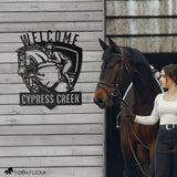 Personalized metal welcome sign for horse barn featuring show jumping horse and rider with custom text ‘Cypress Creek,’ mounted on barn wall beside young woman leading bay horse