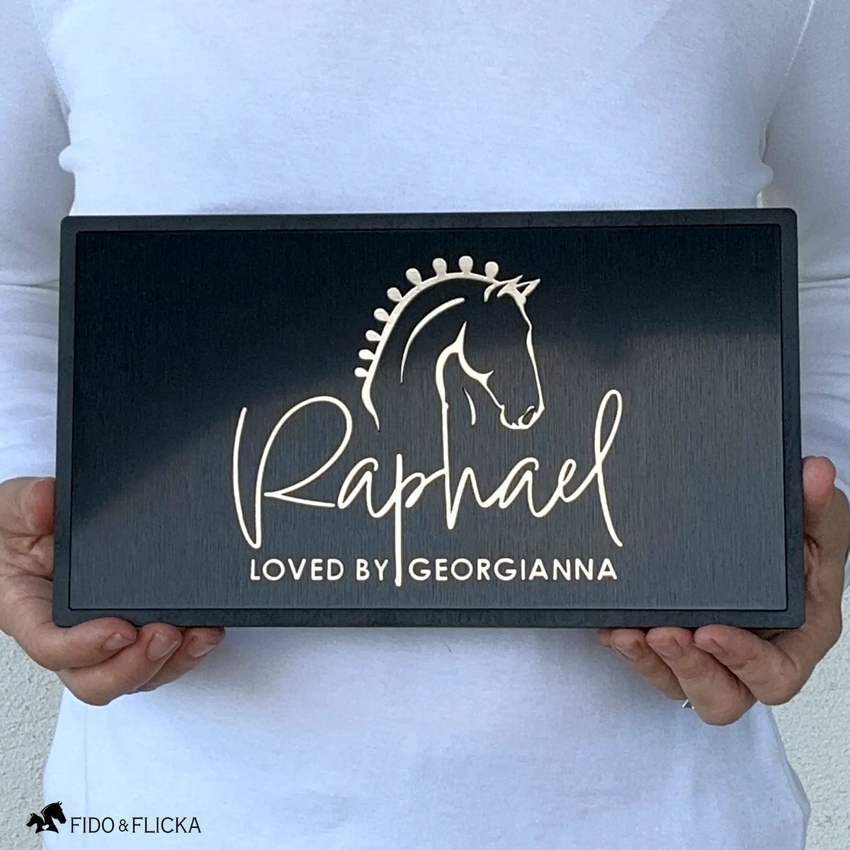 Person holding personalized horse stall nameplate with raised brushed gold acrylic show horse portrait and flowing script name Raphael with loved by Georgianna secondary text on brushed black acrylic base and sealed black wood frame by Fido and Flicka