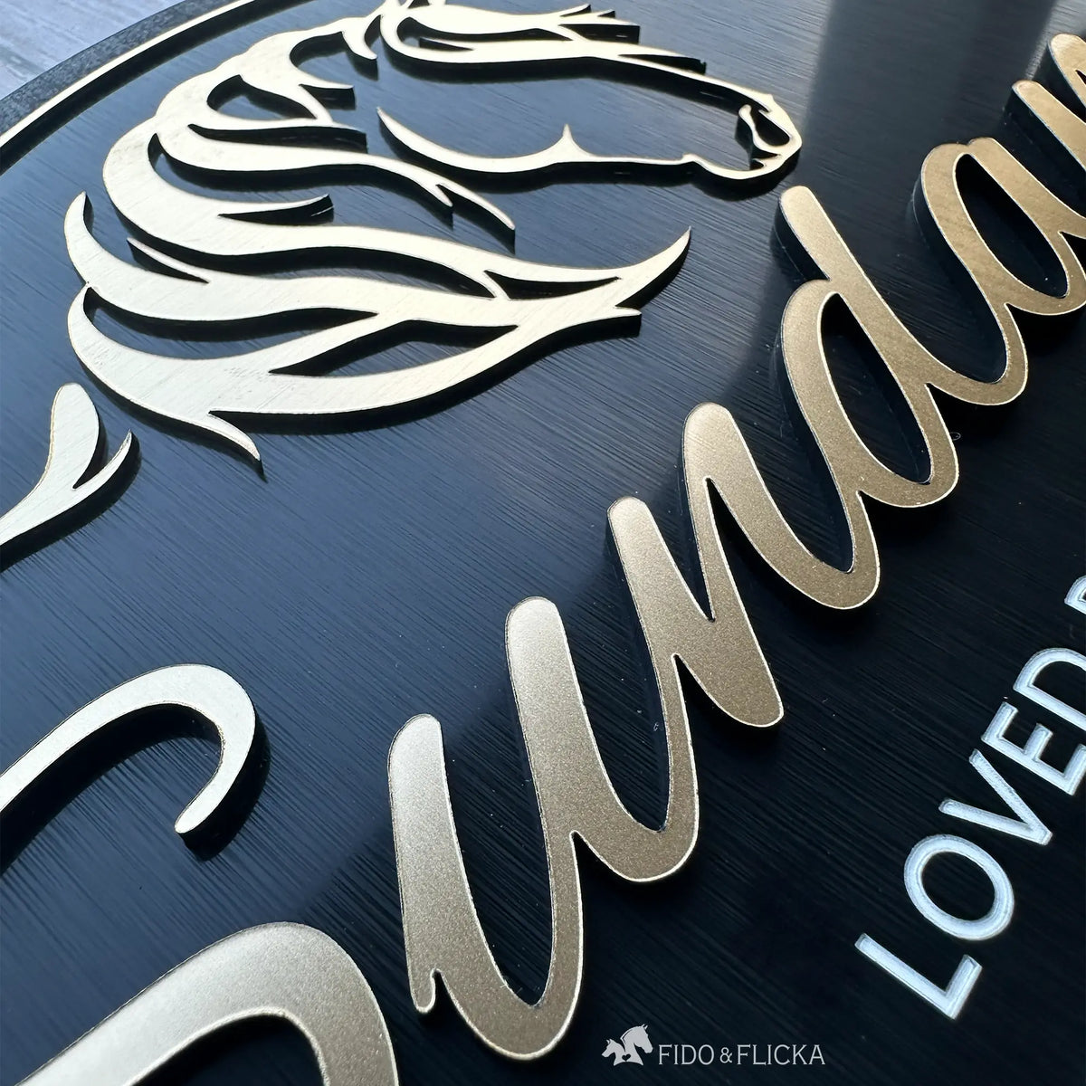 Close-up view of a custom horse stall sign showing layered brushed gold acrylic horse artwork and raised personalized lettering on a black acrylic plaque.