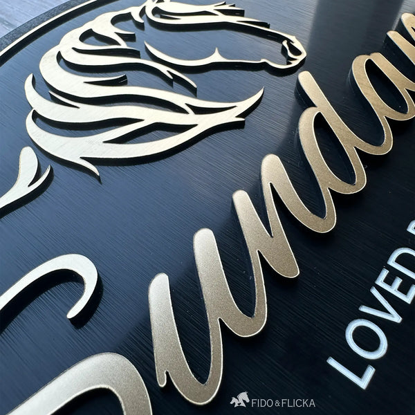 Close-up view of a custom horse stall sign showing layered brushed gold acrylic horse artwork and raised personalized lettering on a black acrylic plaque.