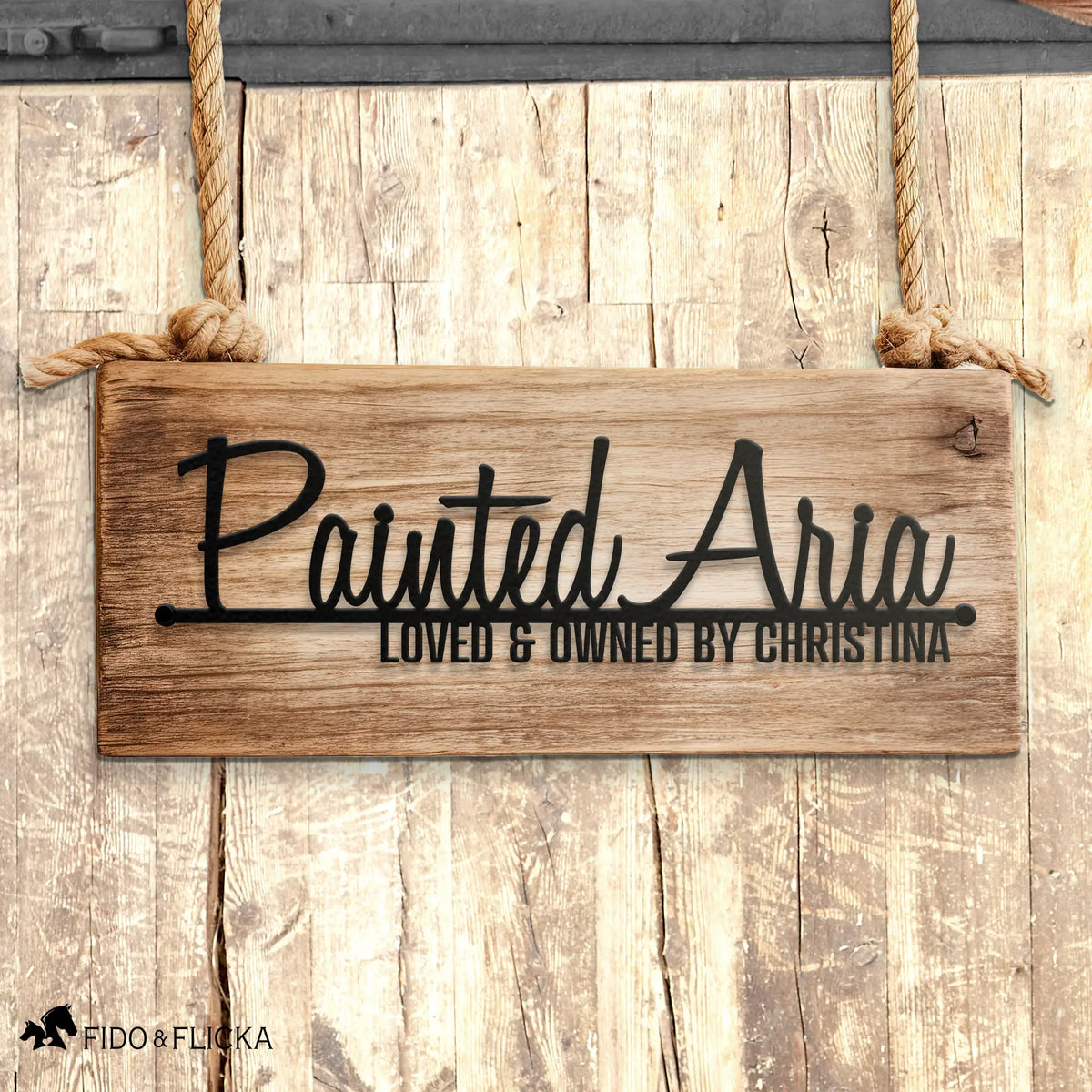 Personalized horse name stall sign reading 'Painted Aria – Loved & Owned by Christina' in black metal on rustic wooden panel, custom barn décor, hanging on horse stall