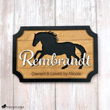 Personalized layered wood horse stall sign featuring a galloping horse silhouette, the name ‘Rembrandt’ in raised white script, and the engraving ‘Owned & Loved by Nicole,’ photographed against a light wood background.
