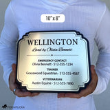 Person holding a 10 × 8-inch brushed gold acrylic horse stall sign with a decorative black border, personalized for Wellington with owner, trainer, and veterinarian contact details.