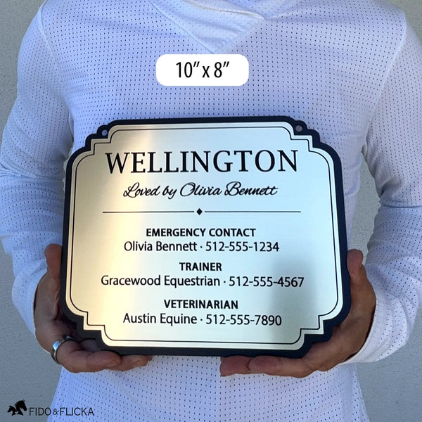 Person holding a 10 × 8-inch brushed gold acrylic horse stall sign with a decorative black border, personalized for Wellington with owner, trainer, and veterinarian contact details.
