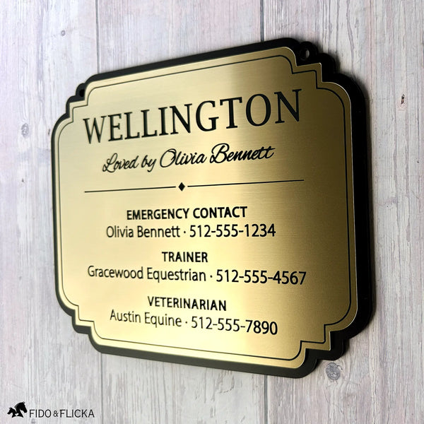 Angled view of Wellington’s personalized brushed gold acrylic stall card sign against whitewashed wood, showing black lettering, a diamond divider, and three emergency contact sections.