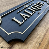 Angled close-up of a black and gold layered wood horse stall name plate reading ‘Lalique,’ showing raised gold lettering, a decorative gold accent, and a gold border on a black painted wood base, photographed on a natural wood surface.