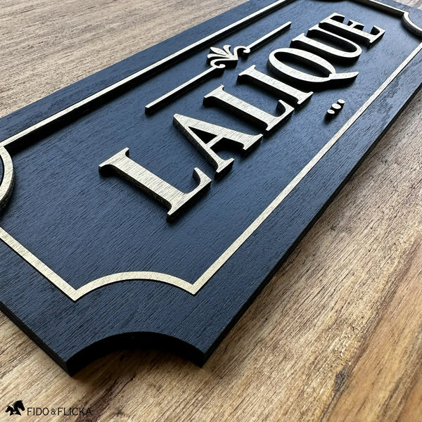 Angled close-up of a black and gold layered wood horse stall name plate reading ‘Lalique,’ showing raised gold lettering, a decorative gold accent, and a gold border on a black painted wood base, photographed on a natural wood surface.