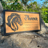 Custom wooden horse stall sign made from Baltic birch with layered black horse head design. Engraved text reads “Phoenix, Owned & Loved by Sheila Jackson.” Personalized barn name plate displayed outdoors on stone with trees in background.