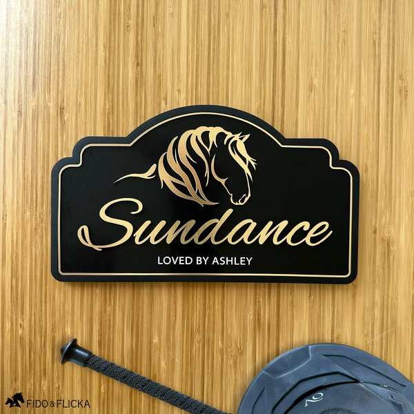 Personalized horse stall sign mounted on a stall door, featuring brushed gold acrylic horse artwork, custom horse name, and engraved text on a black acrylic plaque.
