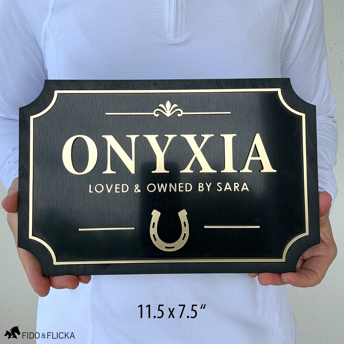Person holding personalized horse stall plaque 11.5 by 7.5 inches showing raised brushed gold acrylic letters spelling Onyxia loved and owned by Sara with horseshoe icon and decorative flourish on brushed black acrylic and sealed black wood frame by Fido and Flicka