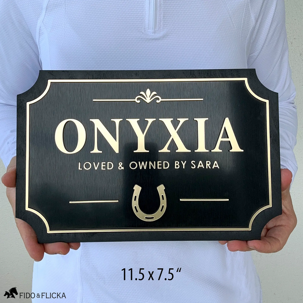 Person holding personalized horse stall plaque 11.5 by 7.5 inches showing raised brushed gold acrylic letters spelling Onyxia loved and owned by Sara with horseshoe icon and decorative flourish on brushed black acrylic and sealed black wood frame by Fido and Flicka