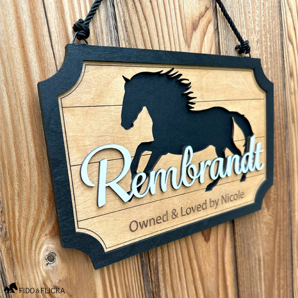 Personalized horse stall sign hanging by a black rope on a wooden barn wall, featuring a running horse silhouette and the raised script name ‘Rembrandt’ above the engraved owner line.