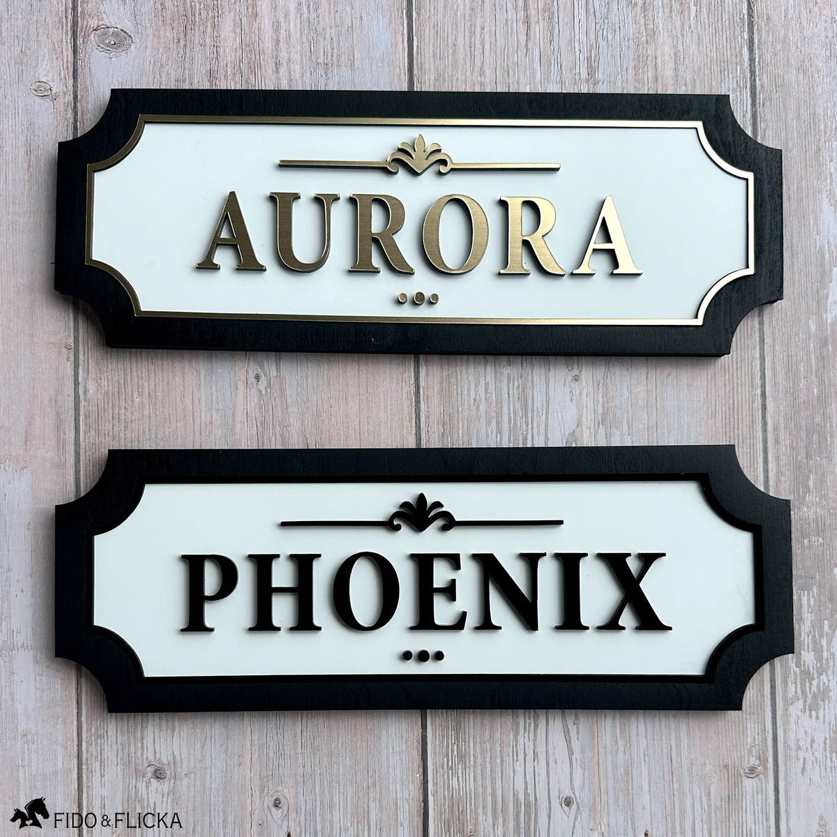 Two custom horse stall name plates displayed together, featuring elegant raised lettering with white acrylic backgrounds and black wood frames in gold and black finishes.
