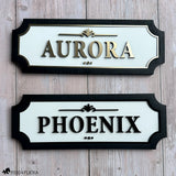 Two custom horse stall name plates displayed together, featuring elegant raised lettering with white acrylic backgrounds and black wood frames in gold and black finishes.