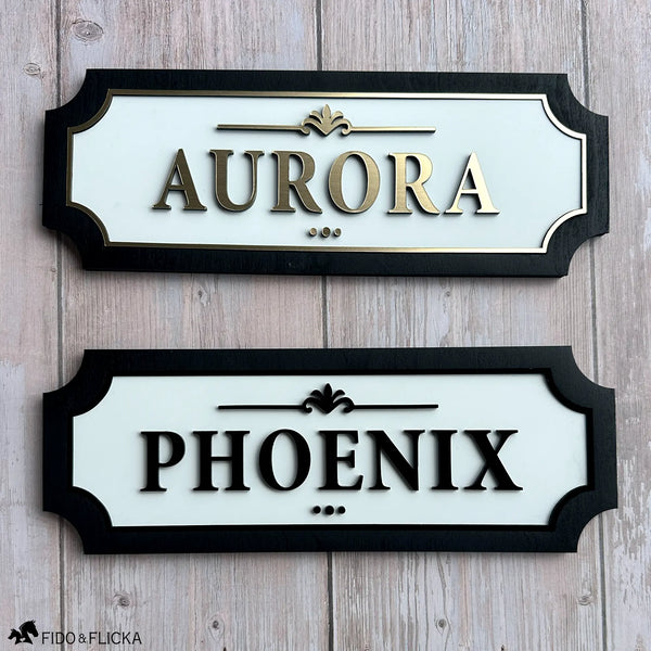 Two custom horse stall name plates displayed together, featuring elegant raised lettering with white acrylic backgrounds and black wood frames in gold and black finishes.