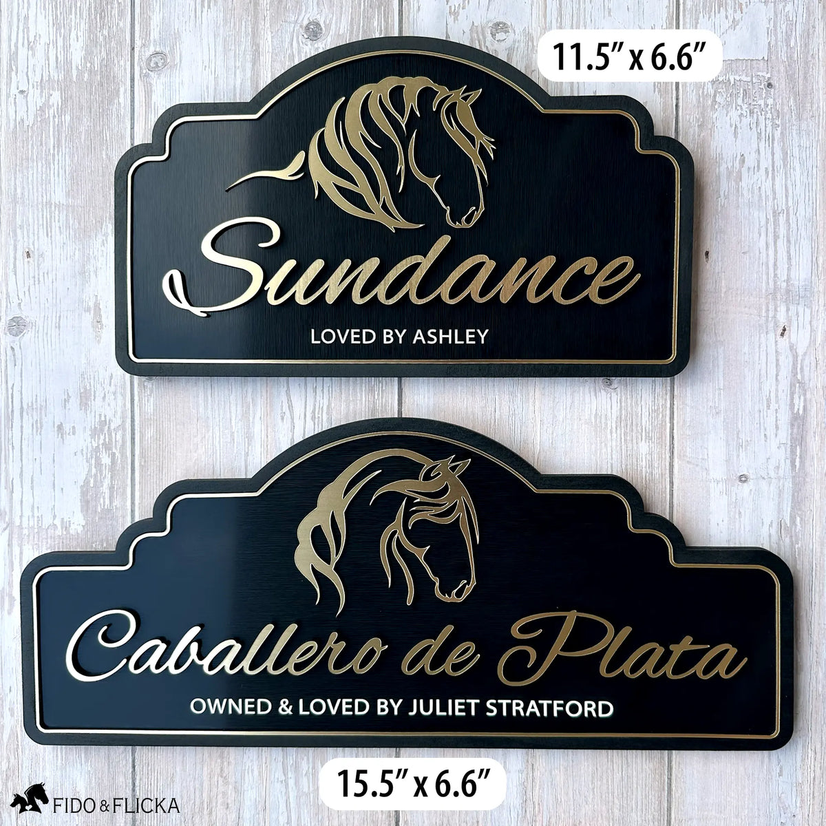 Two personalized horse stall signs in brushed gold on black, shown side by side against a whitewashed wood background. The smaller 11.5-inch by 6.6-inch sign reads "Sundance, Loved by Ashley" with a flowing mane horse silhouette. The larger 15.5-inch by 6.6-inch sign reads "Caballero de Plata, Owned and Loved by Juliet Stratford" with an elegant horse portrait silhouette. Both signs feature raised gold acrylic lettering, a gold border, and an arch-top black wood frame.