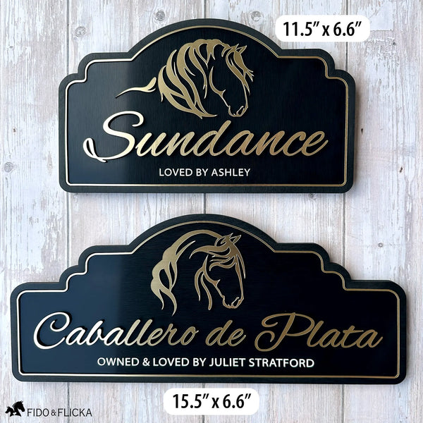 Two personalized horse stall signs in brushed gold on black, shown side by side against a whitewashed wood background. The smaller 11.5-inch by 6.6-inch sign reads "Sundance, Loved by Ashley" with a flowing mane horse silhouette. The larger 15.5-inch by 6.6-inch sign reads "Caballero de Plata, Owned and Loved by Juliet Stratford" with an elegant horse portrait silhouette. Both signs feature raised gold acrylic lettering, a gold border, and an arch-top black wood frame.