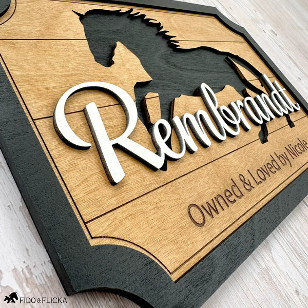 Close-up angled photo showing the layered wood details of a custom horse stall sign with the name ‘Rembrandt’ in raised script over a galloping horse silhouette