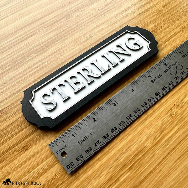White acrylic and wood tack room nameplate with raised brushed silver lettering reading "Sterling" and a scalloped black border, shown next to a metal ruler measuring about 6.5 inches long.