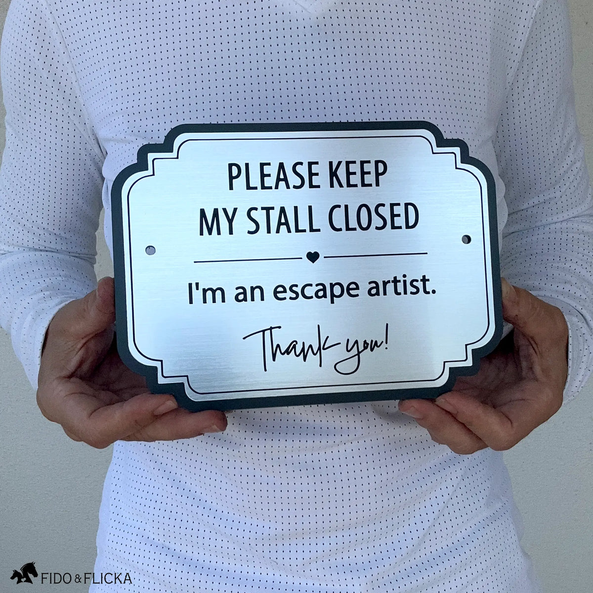 Brushed silver horse stall warning sign reading “Please Keep My Stall Closed, I’m an escape artist,” shown held in two hands for scale.