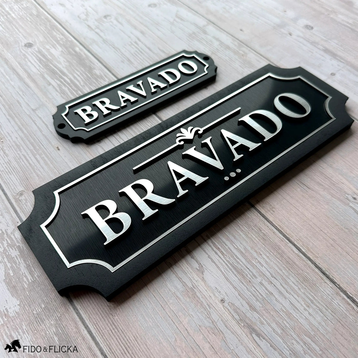 Two matching black acrylic and wood nameplates with silver lettering reading "Bravado" — a smaller tack room plate beside a larger stall nameplate — shown together on weathered wood to show how they coordinate.