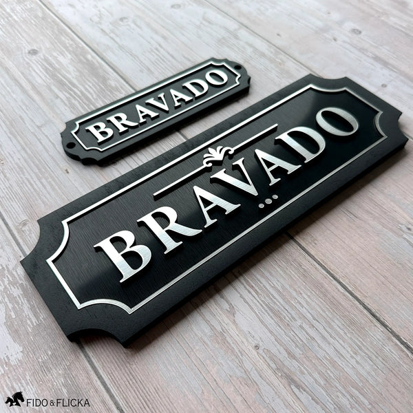 Two matching black acrylic and wood nameplates with silver lettering reading "Bravado" — a smaller tack room plate beside a larger stall nameplate — shown together on weathered wood to show how they coordinate.
