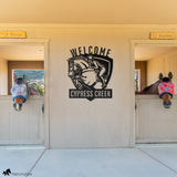 Custom metal horse barn welcome sign with show jumping horse and rider design and personalized text ‘Cypress Creek,’ displayed between two horse stalls with fly-masked horses.